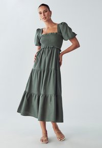 Zelena maksi haljina s nabranoj busti, kratkim puf rukavima i volanima na rubu. Uparena s minimalističkim ravnim sandalama.
