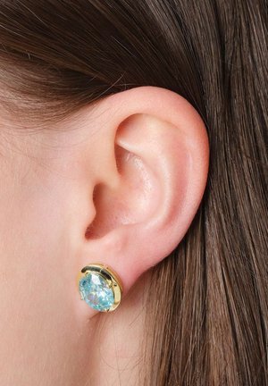 Nahaufnahme eines menschlichen Ohrs mit glattem braunem Haar, das einen goldenen Stecker mit einem großen, runden, hellblauen Edelstein trägt.