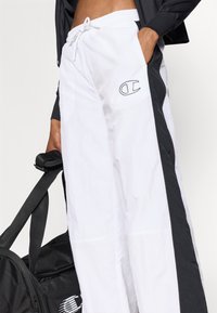 Bílé kalhoty se stažením v pase a černými bočními pruhy a logem, vyrobené z lehkého materiálu, s uvolněným střihem a bočními kapsami.