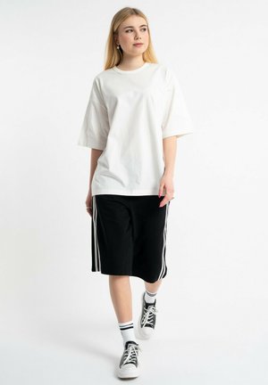 Młoda kobieta o długich blond włosach, ubrana w białą, oversize koszulkę, czarne spodenki do kolan z białymi paskami, białe skarpety i czarne sneakersy.