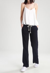White sleeveless top with a flowing design and a rounded hem, paired with dark navy drawstring pants and white sneakers.