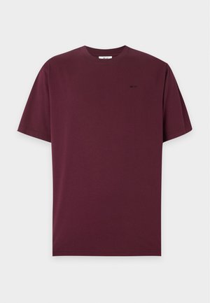 En ung mand iført en mørk burgundy t-shirt, løse sorte bukser, hvide sneakers og blåtonede solbriller står foran en ensfarvet baggrund.