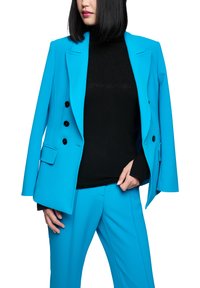 Frau trägt einen leuchtend blauen Anzug mit schwarzem Rollkragenpullover, eine Hand passt die Jacke an, schwarze Bobfrisur, vor weißem Hintergrund.