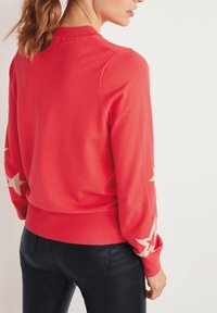 Roter Pullover mit langen Ärmeln, geripptem Ausschnitt und Bündchen, mit cremefarbenen Sternmustern auf den Ärmeln. Trägt dazu schwarze Hosen.