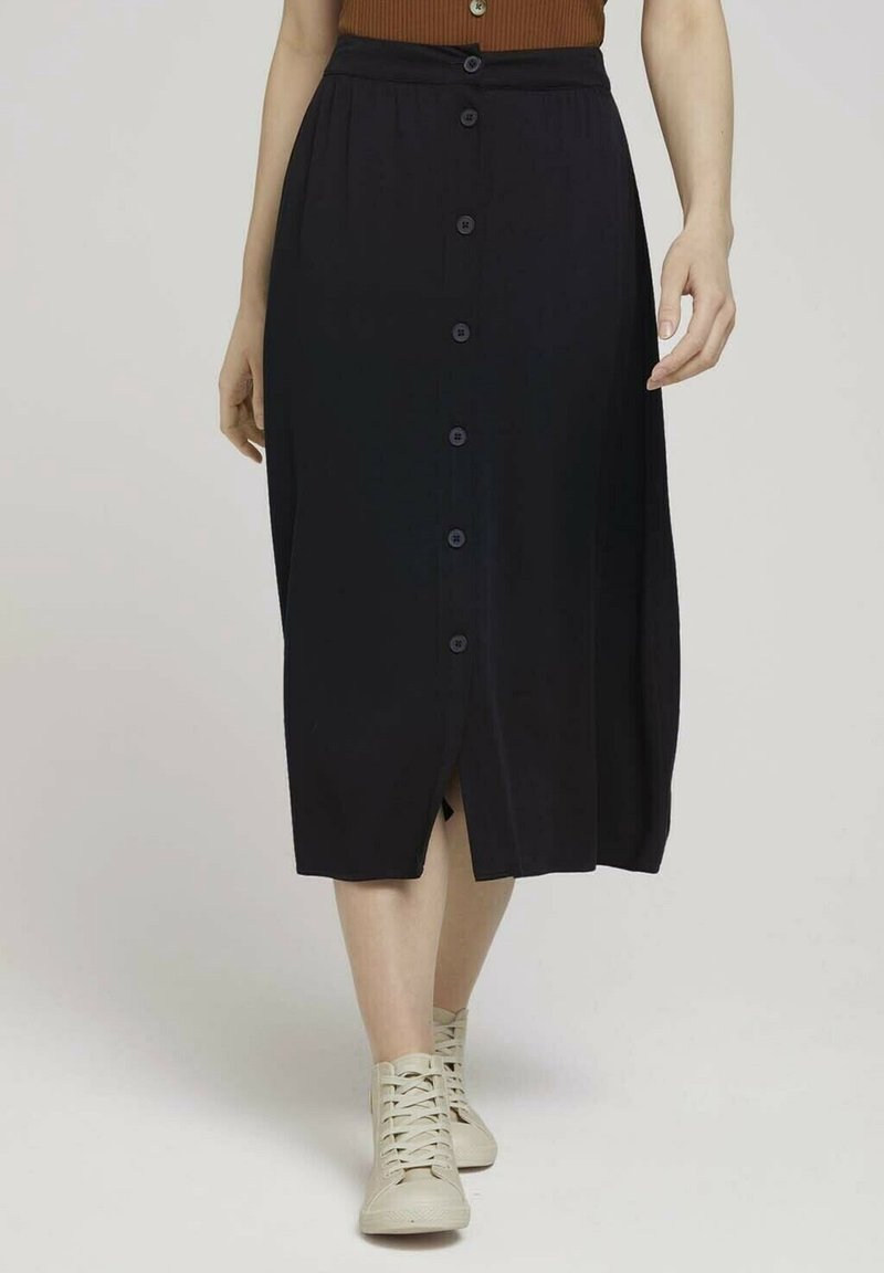 Femme portant une jupe midi noire à boutons sur le devant, un haut marron et des baskets montantes beige, marchant sur un fond clair uni.