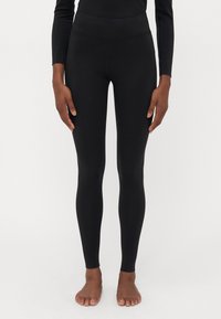 Asmuo stovintis basomis, vilkintis prigludusias juodas leggins ir ilgas rankovines juodas palaidinę prieš vienodą šviesią foną.