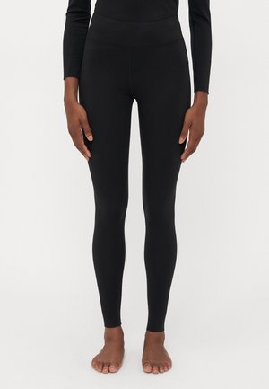Asmuo stovintis basomis, vilkintis prigludusias juodas leggins ir ilgas rankovines juodas palaidinę prieš vienodą šviesią foną.