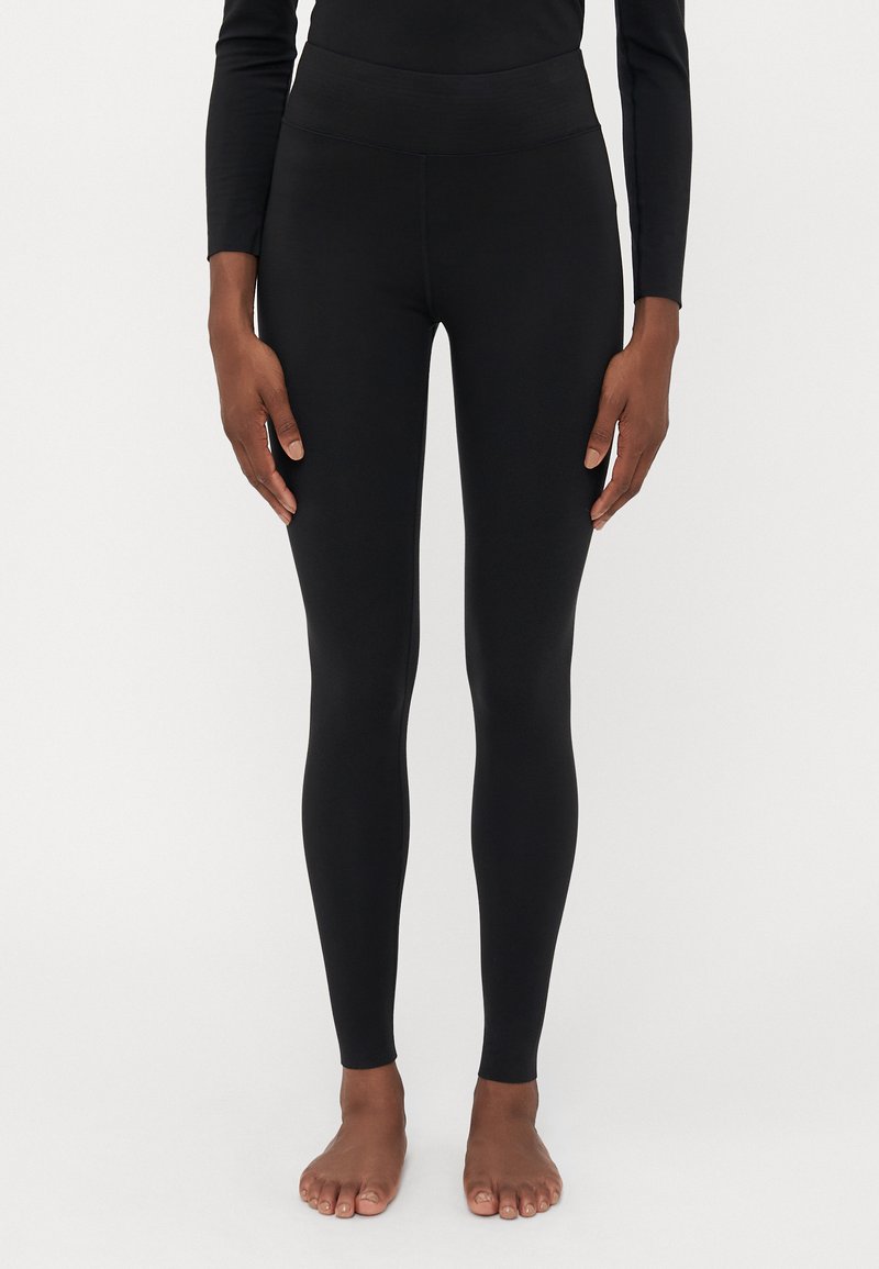Person som står barfota iklädd åtsittande svarta leggings och en långärmad svart topp mot en enkel ljus bakgrund.