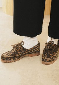 Scarpe in suede con stampa leopardata e suole in gomma marrone, design con lacci e finitura testurizzata, abbinate a calzini bianchi e pantaloni neri.