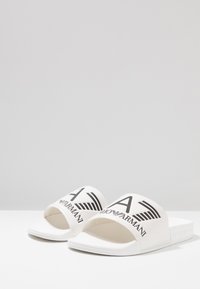 Valkoiset liukkumäntysandaalit, jotka on valmistettu kumista. Ne sisältävät leveän hihnan, jossa on musta logo-teksti ja kotka-grafiikka. Tasapohja, jossa on teksturoitua yksityiskohtaa.