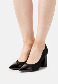 Chaussures à talons hauts noires avec dessus en daim et bout pointu en cuir verni. Le design à talon carré offre stabilité et un look moderne.