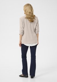 Beige, lichte blouse met opgerolde mouwen, gelaagd over een wit shirt; gecombineerd met donkerblauwe, uitlopende jeans en zwarte ballerina's. Achteraanzicht.
