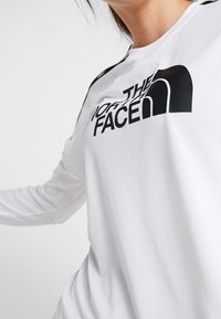 Vit långärmad skjorta i smidigt tyg med en stor svart "The North Face"-logotyp på framsidan och svarta detaljer på axlarna.