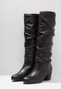 Bottes noires en cuir montant jusqu'au genou, au design froncé, avec un bout rond et un talon épais. Texture lisse et style minimaliste.