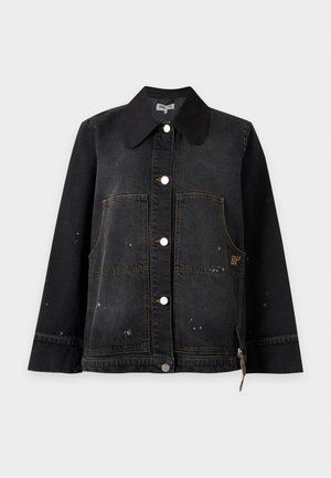 Femme portant une veste en jean noire ample, un pantalon blanc à jambes larges et des chaussures marron pointues, debout sur un fond uni.