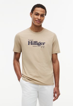 Ung mand iført beige Tommy Hilfiger t-shirt med logo og hvide bukser, stående med den ene hånd i lommen mod en hvid baggrund.