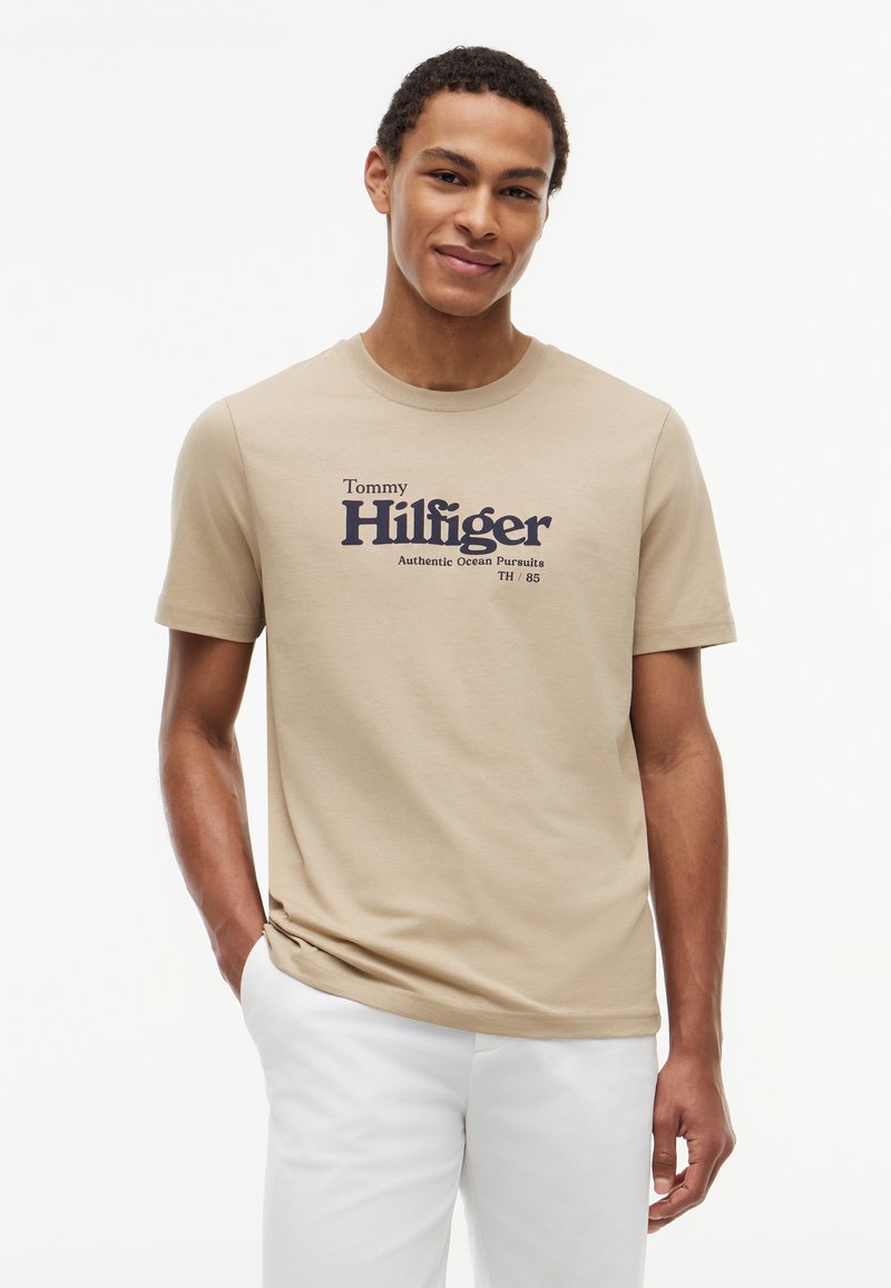 Jeune homme portant un t-shirt beige Tommy Hilfiger avec le logo et un pantalon blanc, debout avec une main dans la poche sur fond blanc.