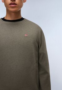 Olivgrüner Strickpullover mit schwarzem Kragen; verfügt über ein kleines rot-weißes Flaggenemblem auf der linken Brust; gerippte Textur.