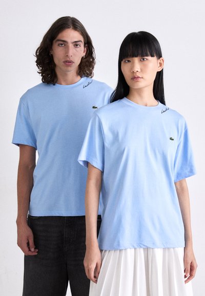 Двама души, облечени с светлосини тениски Lacoste, единият с тъмни дънки, а другият с бяла плисирана пола, стоят на фона на прост фон.