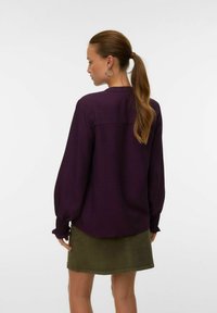 Donna con una lunga coda di cavallo che indossa orecchini a cerchio, una blusa larga di un viola scuro con polsini arricciati e una gonna verde oliva, girata di spalle.