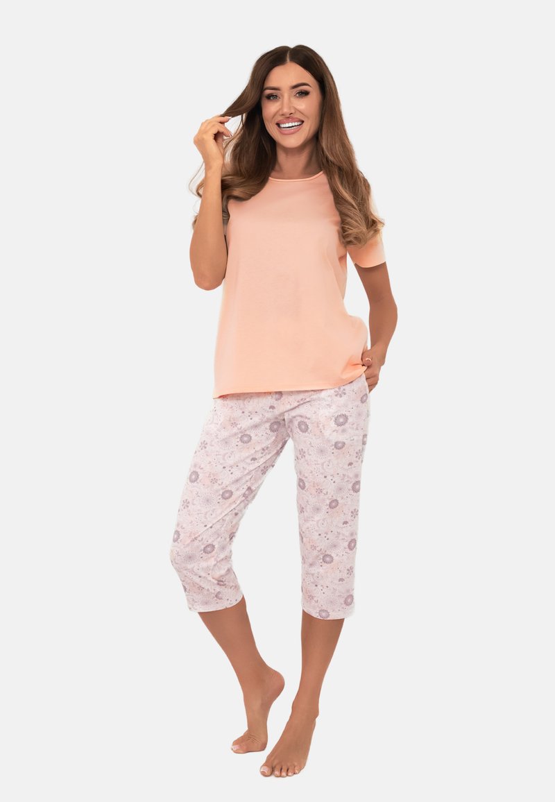 Kobieta uśmiechająca się, ubrana w brzoskwiniowy top z krótkim rękawem i białe spodnie capri w różowy kwiatowy wzór, stojąca boso na białym tle.