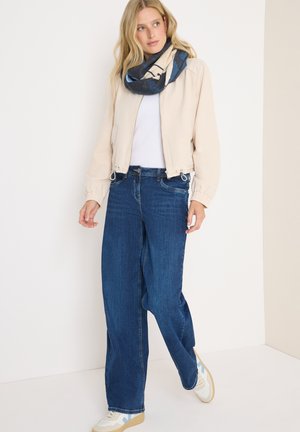 Giacca bomber beige, t-shirt bianca, jeans larghi blu e sciarpa di blu con motivi. Le sneakers presentano colori chiari e accenti sottili.