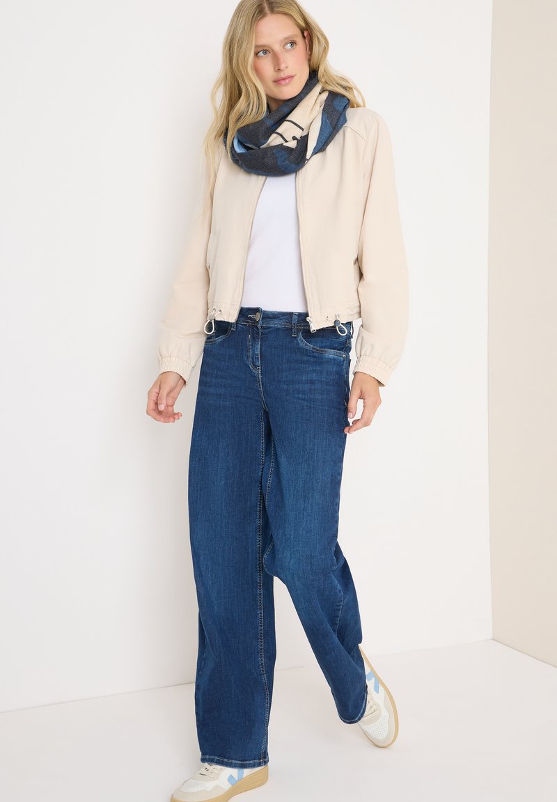 Beige Bomberjacke, weißes T-Shirt, blaue weit geschnittene Jeans und ein gemusterter blauer Schal. Die Sneakers haben helle Farben und subtile Akzente.
