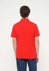 Piros galléros póló lapos nyakkal és rövid ujjal. Sima anyagból készült, egyenes szegéllyel. Kék farmernadrággal viselve.