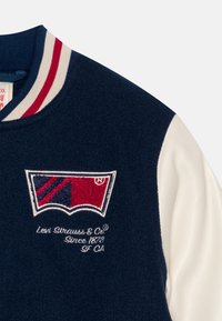 Námořnická vlněná baseballová bunda s bílými koženými rukávy. Na bundě je vyšitý červený a modrý logo a bílý, červeně pruhovaný žebrovaný límec.