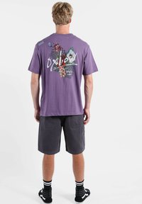 T-shirt violet en coton avec impression graphique florale et texte « O'Neill » au dos, associé à un short cargo gris et des chaussettes rayées noir et blanc.
