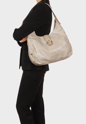 Donna con abito nero che porta una grande borsa a tracolla in pelle beige con una fibbia circolare dorata e un marchio discreto.