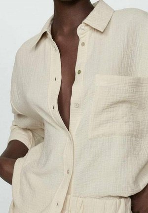 Person iført en åben, tekstureret beige skjorte med brystlomme og opkørte ærmer, kombineret med matchende bukser med høj talje.