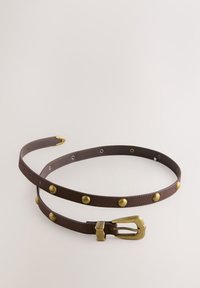 Ceinture en cuir marron avec clous ronds en laiton et boucle en laiton, posée enroulée sur un fond clair et neutre.