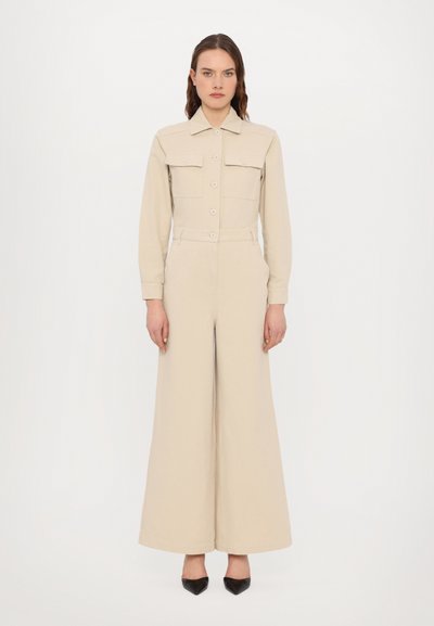 Beige jumpsuit with long sleeves, collar, two front pockets, belted waist, and wide leg design. Smooth fabric with a minimalistic look.