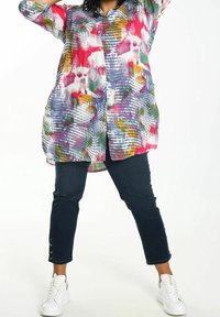 Person, der ein bunt gemustertes langes Hemd, dunkle Jeans mit Knopfdetails am Saum und weiße Sneaker trägt, steht mit gespreizten Beinen.