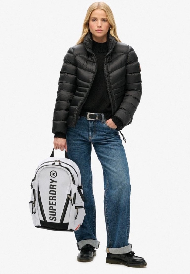 Doudoune noire, pull en laine noir, jeans évasés bleu clair, et un sac à dos blanc avec un logo noir. Chaussures noires avec une finition brillante.