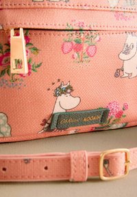 Rožinė drobės rankinė su Moomin personažais ir gėlių raštais. Aukso detalės ir žalia etiketė su užrašu "Cath Kidston Moomin" prideda elegancijos.