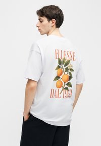 Νεαρός άνδρας που κοιτάζει προς τα πλάγια φοράει λευκό, φαρδύ T-shirt με εκτύπωση πορτοκαλιού και πράσινων φύλλων, και το κείμενο "ELLESSE DAL 1959" στην πλάτη.