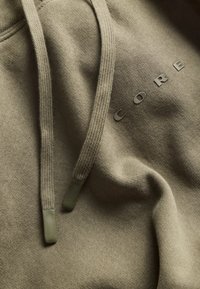 Olive green hoodie with a soft texture, featuring a logo reading "CORE" in a subtle, embossed style. Drawstrings are textured.