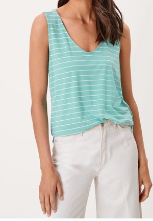 Kobieta w błękitno-turkusowej, bezrękawnikowej bluzce w paski i białych spodniach, stojąca z lewą ręką swobodnie opuszczoną wzdłuż ciała.