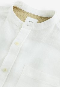 Chemise en lin blanc avec un col montant, fermeture à boutons et une poche poitrine. L'intérieur du col présente un tissu à motif léger.