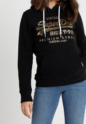 Kobieta w czarnej bluzie z kapturem z złotym, vintage logo „SuperDry” i białymi troczkami, w połączeniu z niebieskimi dżinsami, stojąca na jednolitym tle.