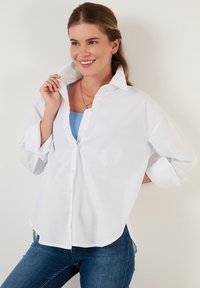 White button-up shirt with a relaxed fit, rolled cuffs, and a rounded hem, layered over a blue top, paired with blue jeans.