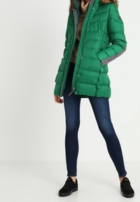 Grön vadderad jacka med grå detaljer, dragkedjestängning och huva. Bärs över en beige topp, tillsammans med blå jeans och svarta sneakers.