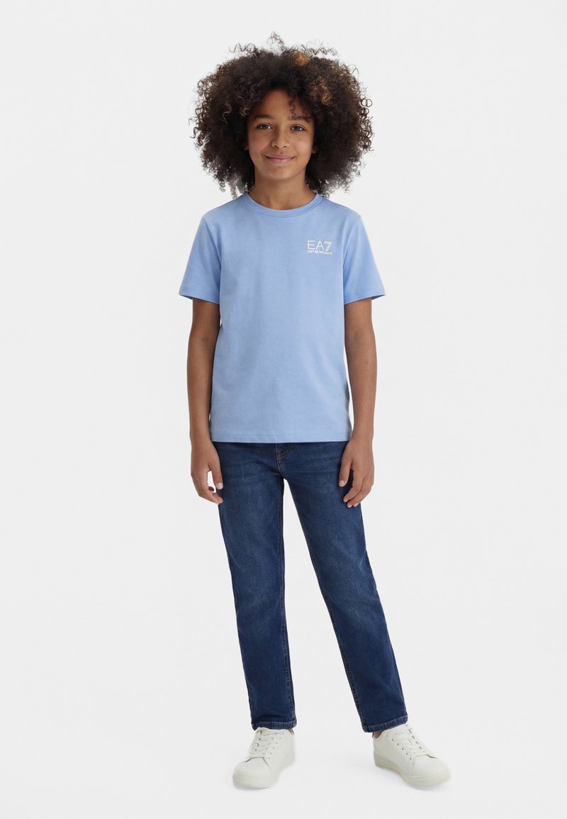 Enfant aux cheveux bouclés portant un t-shirt EA7 bleu clair, un jean bleu foncé et des baskets blanches, debout devant un fond blanc uni.