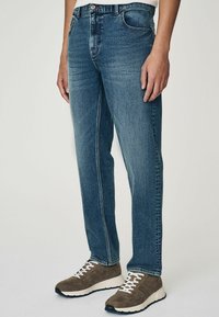 Denim jeans in een middelblauwe kleur met een rechte snit. Voorzien van vijf zakken en contrasterende stiksels. Gecombineerd met bruin suède sneakers.