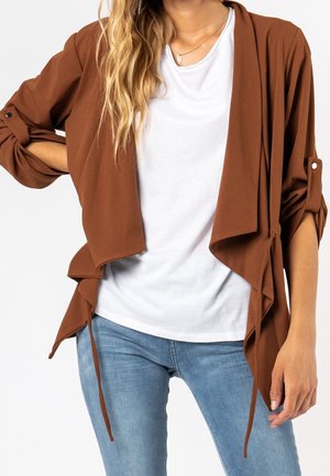 Femme portant un jean bleu clair, un t-shirt blanc et un cardigan marron ample ouvert sur le devant, avec des manches retroussées et des détails à nouer.