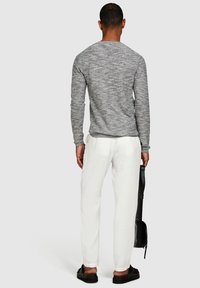 Grå texturerad långärmad skjorta, vita linbyxor och svarta sandaler. Modellen håller en svart väska, sedd bakifrån mot en grå bakgrund.