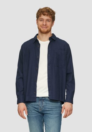 Navyblaues Hemd mit Knopfverschluss aus leichtem Stoff, mit einer Brusttasche, über einem weißen T-Shirt getragen, kombiniert mit hellblauen Jeans.