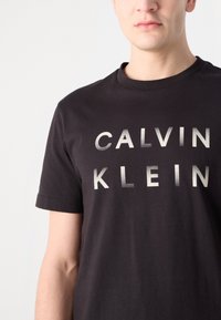 Tricou negru din bumbac cu text metalic gri "CALVIN KLEIN" în litere mari, îngroșate. Mâneci scurte și decolteu rotund.